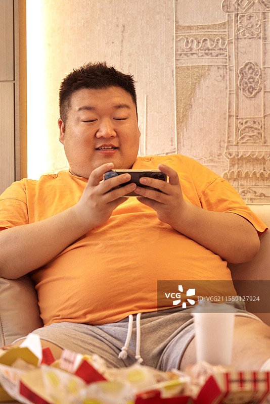 肥胖的青年男人躺在沙发上玩手机图片素材