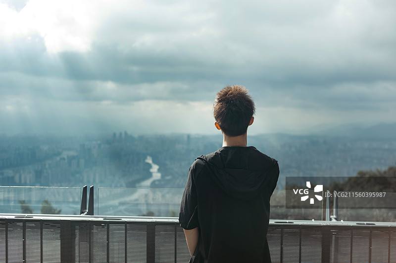 一位青年站在山顶眺望远方的城市天际线，象征自由、探索与旅行精神。图片素材