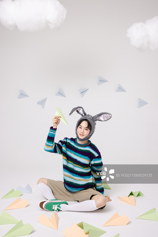 年轻男子戴着兔子帽，坐在纸飞机的包围中。图片素材