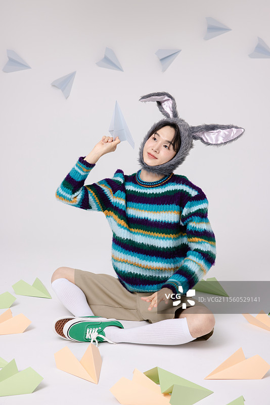 年轻男子戴着兔子帽，坐在纸飞机的包围中。图片素材
