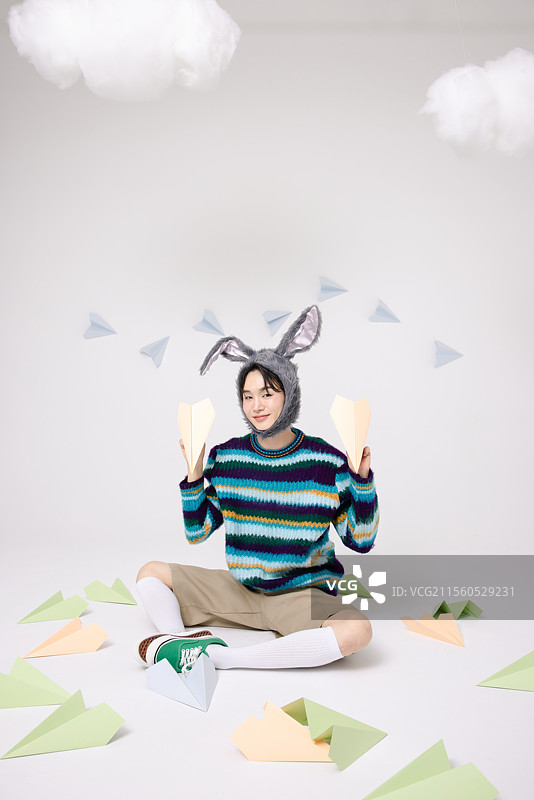 年轻男子戴着兔子帽，坐在纸飞机的包围中。图片素材