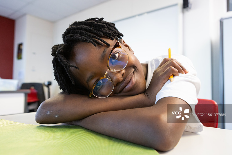 在学校，微笑的非裔美国男孩戴着眼镜，手握铅笔，靠在桌子上休息。图片素材