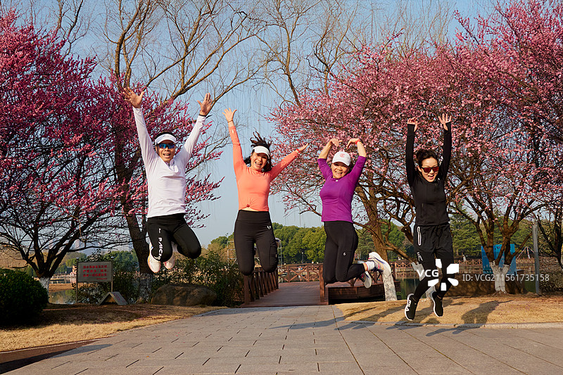四位身着运动装的在红梅开放的公园石板路上跳跃合影，背景融合蓝天绿树与桥梁，洋溢着春日活力与青图片素材