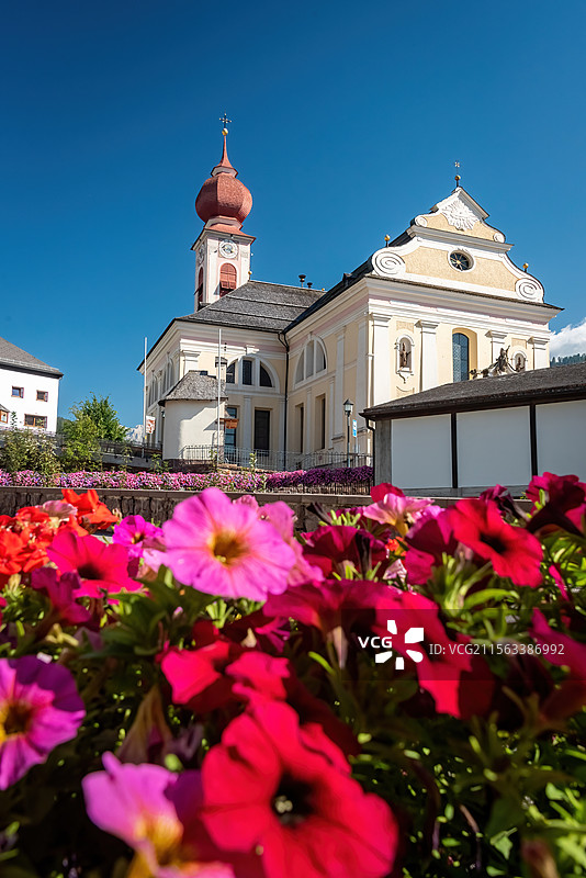 意大利多诺米蒂建筑在晴朗的蓝天下显得格外清新，红色圆顶塔楼和风向标。前景盛开的粉红色和红色矮牵牛花图片素材