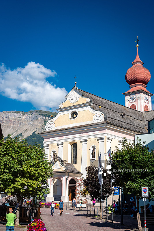 意大利多诺米蒂白云岩宁静的教堂伫立在蓝天和山脉的背景下，红色圆顶塔楼,教堂周围有树木和花坛点缀图片素材