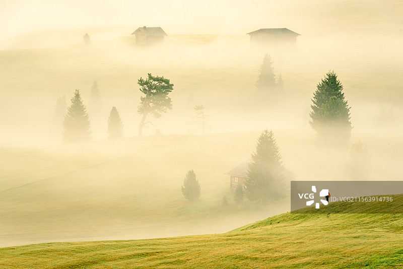 意大利多诺米蒂白云岩宁静的山间，一座山雾中，行者手持登山杖，悠然行走于山坡。远处的木屋与树木若隐若现图片素材