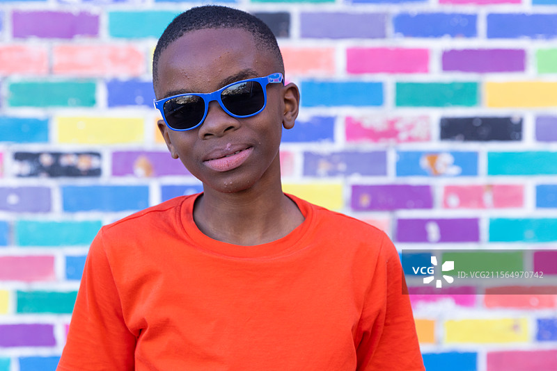 戴着太阳镜的男孩穿着橙色衬衫，站在五彩斑斓的砖墙前图片素材
