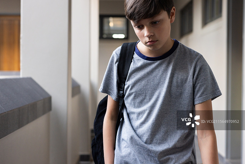 在学校走廊里，一个背着书包的男孩低头走着，显得很伤心。图片素材