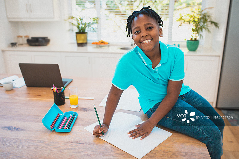 非裔美国男孩坐在家里的厨房岛上，正在用笔记本写字，旁边有一台笔记本电脑，留有空白空间。图片素材