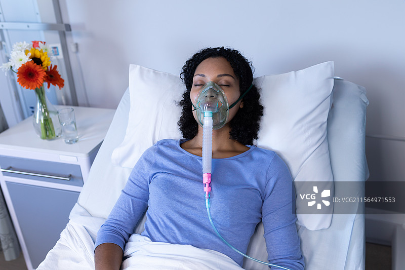 混血年轻女性躺在医院病床上，佩戴氧气面罩，桌上放着鲜花。图片素材
