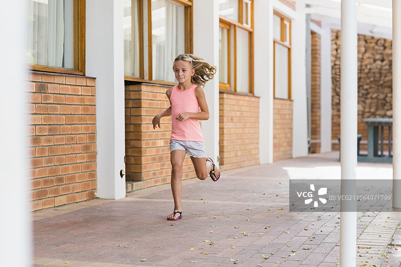 女孩在学校的砖砌走廊上奔跑，走廊有木窗、柱子和树叶。图片素材