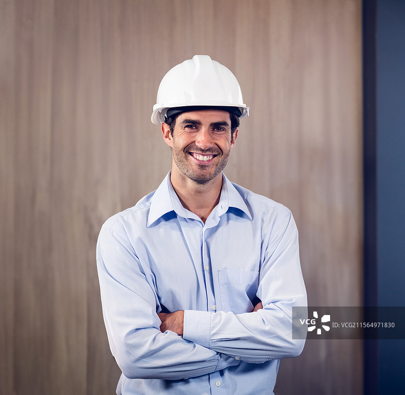 一名男子站在工地办公室，戴着白色安全帽，穿着浅蓝色衬衫，自信地微笑。图片素材