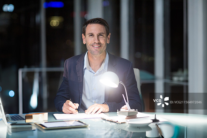 男子在现代办公室的桌子上夜间用笔记本写字，旁边有笔记本电脑和城市灯光。图片素材