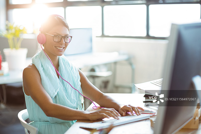 在现代办公室桌前，戴着粉色耳机和眼镜的女性正在键盘上打字图片素材