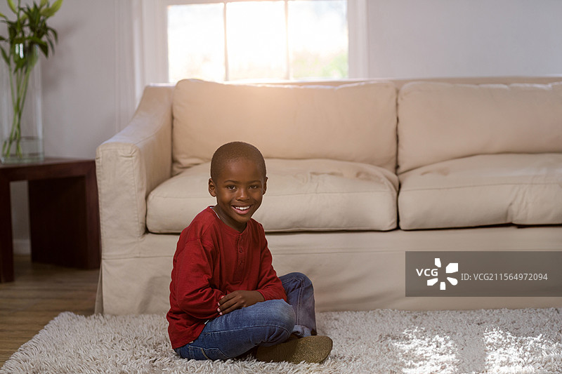 非裔美国儿童盘腿坐在客厅的长毛地毯上，面带微笑，旁边有一盆绿色植物。图片素材