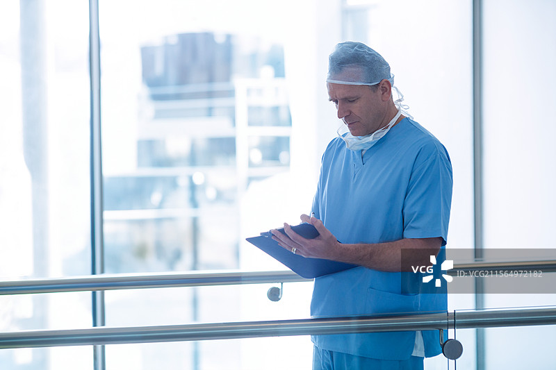 戴着外科口罩的男子在医院走廊的栏杆旁查看夹板，留有空白空间图片素材