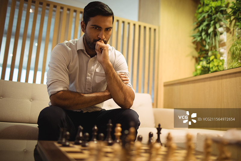 年轻的亚洲男子坐在现代休息室的沙发上，正在分析木制棋盘上的棋子图片素材