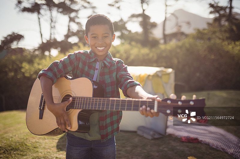 在公园里，一名非裔美国男孩朝着镜头举着木吉他，旁边有一个冷藏箱和格子毯子。图片素材