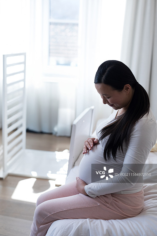 亚洲孕妇坐在婴儿房的床边，轻抚着肚子，旁边是白色婴儿床，留有空白空间。图片素材
