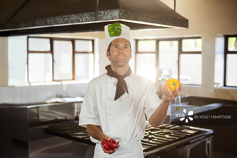 中年男性厨师在商业厨房中抛接三个彩椒，展示色彩丰富的农产品图片素材