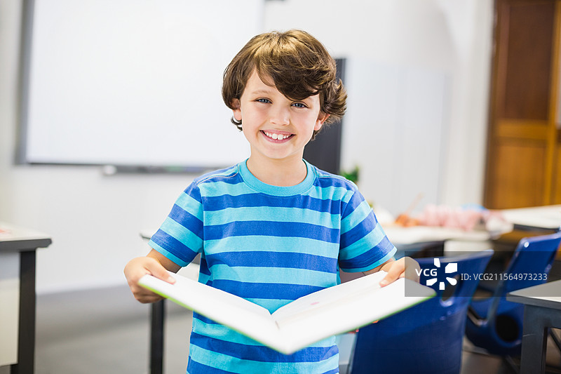 孩子在教室里拿着一本打开的精装书，周围有蓝色塑料椅子和桌子。图片素材