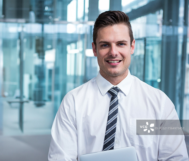 年轻男子站在现代办公室大堂，手持笔记本电脑，展示企业工作空间图片素材