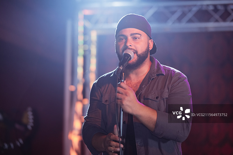 男子在音乐会舞台上握着麦克风架唱歌，背景有灯光架和模糊的吉他琴头。图片素材
