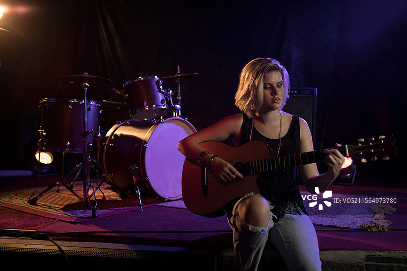 青少年女孩在小型室内舞台上弹奏木吉他，旁边有鼓组和音响，留有空白空间图片素材