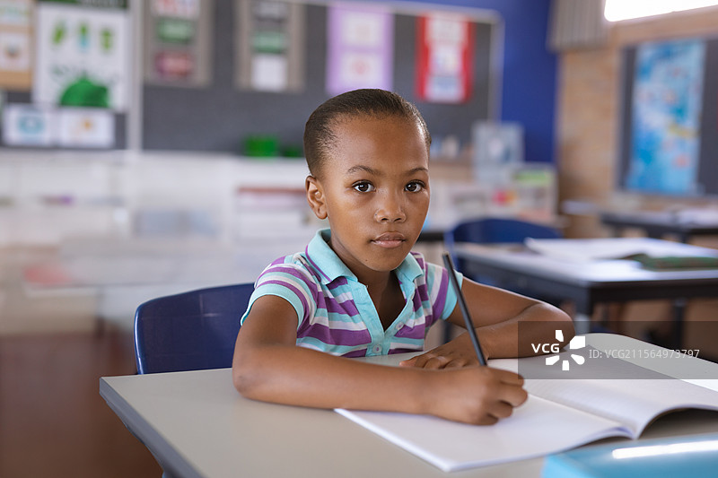 非裔美国儿童在阳光明媚的教室里用削尖的铅笔在桌子上写字，旁边是打开的笔记本。图片素材