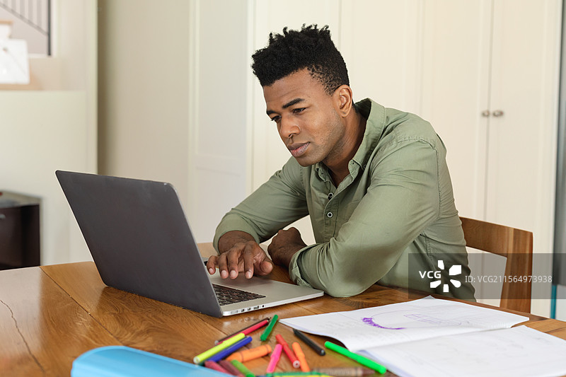 非裔美国男性在家中的木桌上使用笔记本电脑，桌上有草图、马克笔和空白空间图片素材