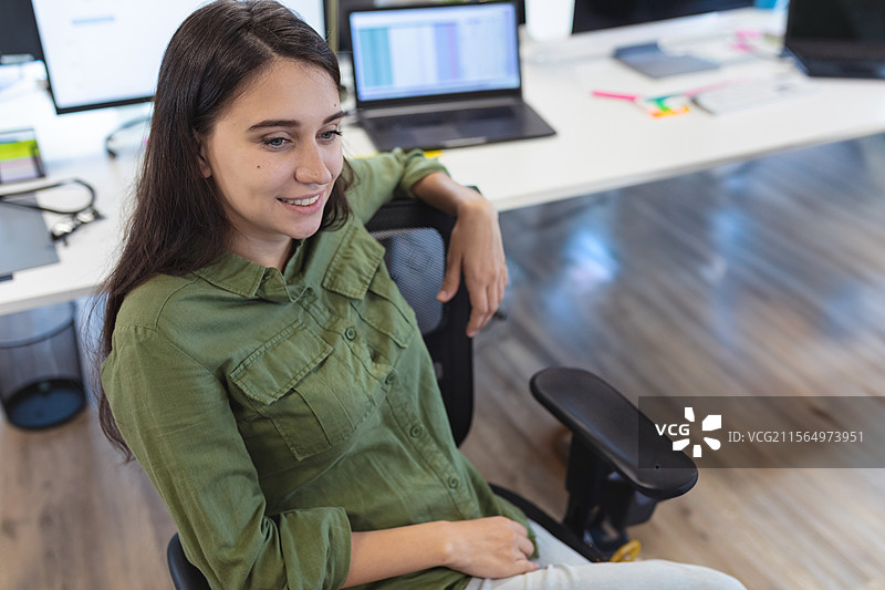 年轻女性靠在办公室桌子的符合人体工程学的椅子上，使用笔记本电脑和显示器。图片素材