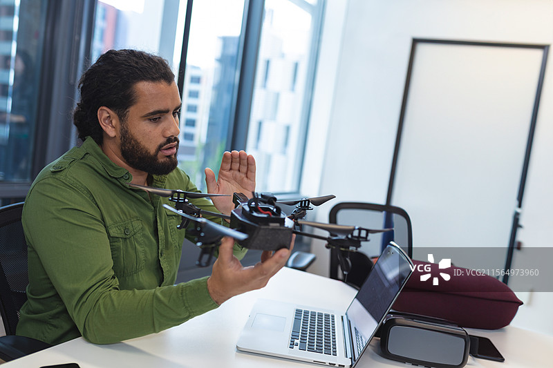 男子在现代办公室桌子旁检查四旋翼无人机，桌上有笔记本电脑和虚拟现实头盔。图片素材