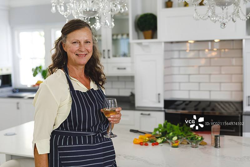 穿围裙的老年女性站在厨房里，手里拿着酒杯、甜椒和刀，留有空白空间。图片素材