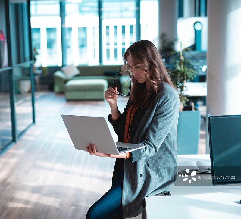 年轻女性站在现代开放式办公室里，手持笔记本电脑并调整眼镜。图片素材