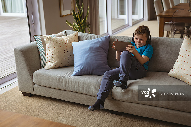 坐在灰色沙发上的学龄男孩，戴着耳机使用平板电脑，客厅内有空白空间。图片素材