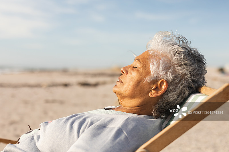 一位老年女性在沙滩上躺在带条纹垫子的躺椅上，享受阳光，旁边留有空白空间。图片素材