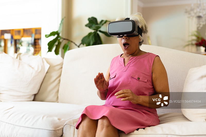 一位非洲裔美国老年女性在客厅沙发上佩戴虚拟现实头盔，惊讶地反应。图片素材