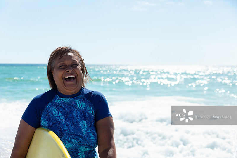 一位穿着蓝色防晒衣、手持黄色冲浪板的老年女性站在海岸线，开心地笑着。图片素材