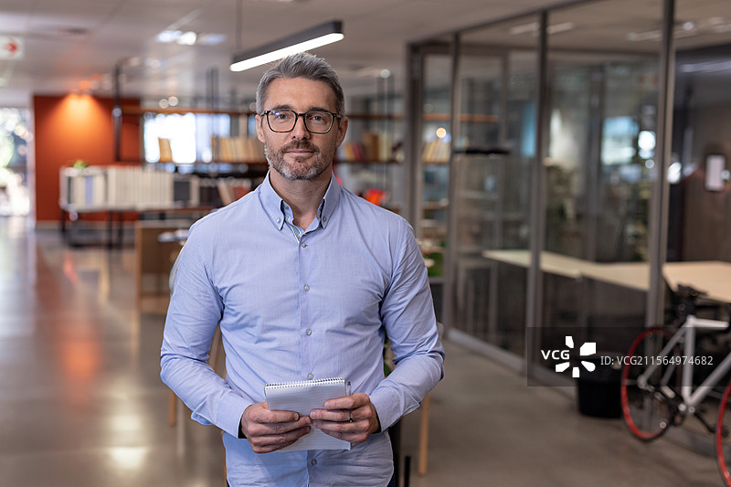 男子在开放式办公室中央手持小型螺旋笔记本，周围有书籍和自行车。图片素材