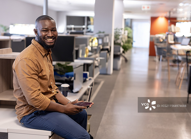 年轻的非裔美国男性坐在开放式办公室的长椅上，手持智能手机，留有空白空间图片素材
