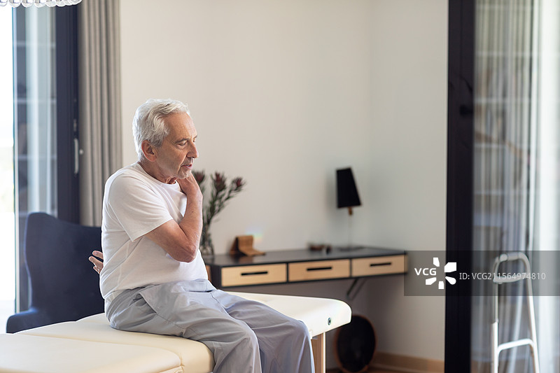 老年男子坐在治疗室的治疗台上，手握着疼痛的脖子，旁边有助行器，留有空白空间。图片素材