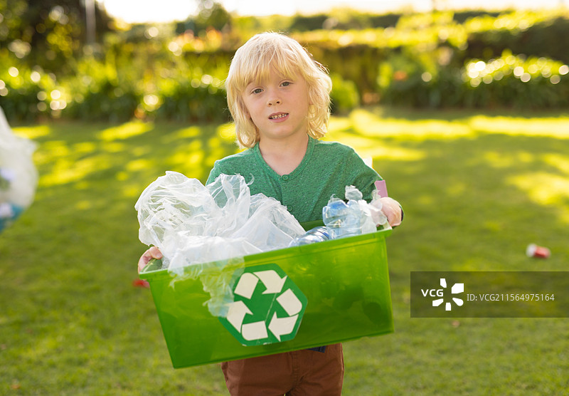 男孩在阳光明媚的后院里拿着绿色回收箱，正在分类塑料瓶和透明袋。图片素材