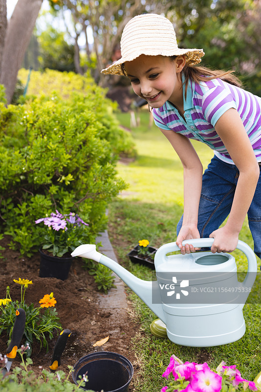 孩子在阳光明媚的后院用蓝色浇水壶和园艺工具给黄色和粉色的花浇水图片素材