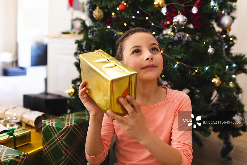 白人女孩坐在客厅的地板上，靠近圣诞树，手里拿着一个金色包装的礼物靠在耳边。图片素材