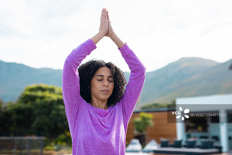 女性在现代露台上做瑜伽祈祷姿势，周围有绿色灌木和山丘。图片素材