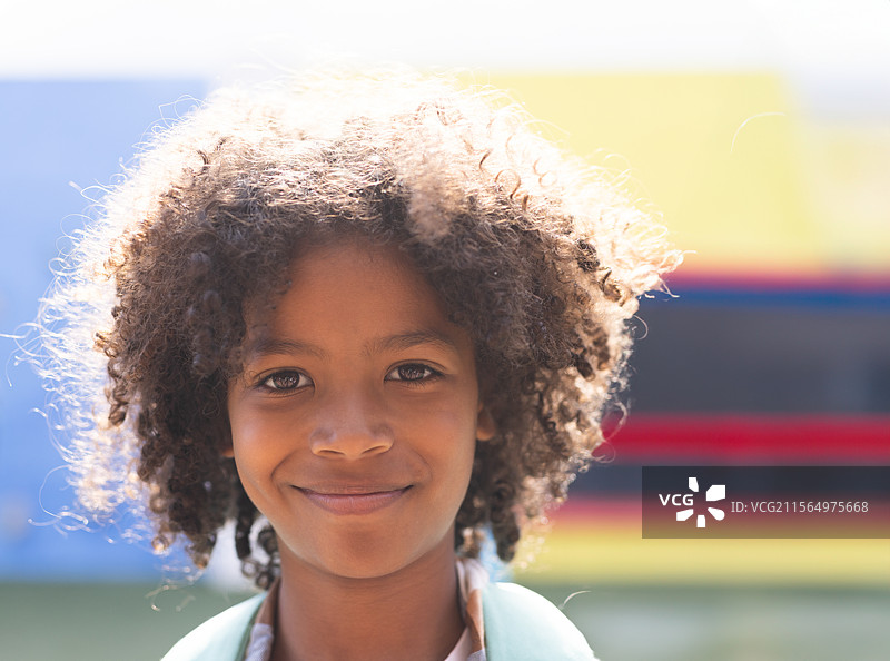 户外一名微笑的小学生站在涂鸦墙前，展现出卷曲的头发和阳光。图片素材