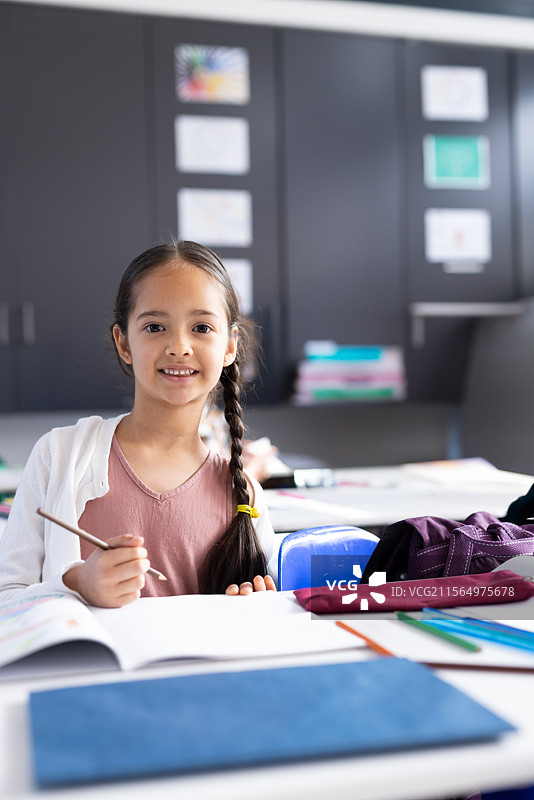 女童坐在学校教室的桌子旁，手握铅笔，旁边有作业本和彩色铅笔。图片素材