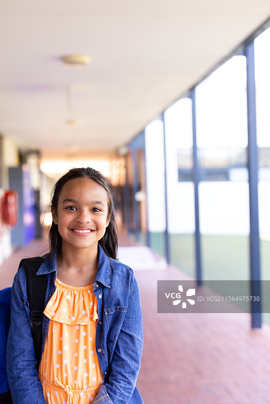 8岁女孩站在有顶的学校走廊里，穿着黄色连衣裙，手里拿着背包。图片素材