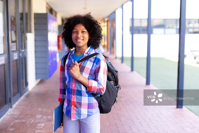 非裔美国男孩在学校走廊里站着，背着黑色背包，手里拿着蓝色文件夹。图片素材