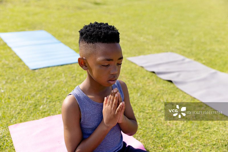 非裔美国男孩跪在粉色瑜伽垫上，在草地公园中闭眼冥想图片素材
